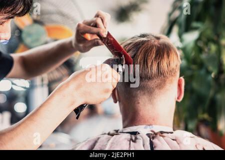 Salone fotografico Lifestyle. Il cliente giovane indossa la maschera facciale, vista posteriore laterale. Regolacapelli elettrico per pettini da lavoro maschio professionale Foto Stock