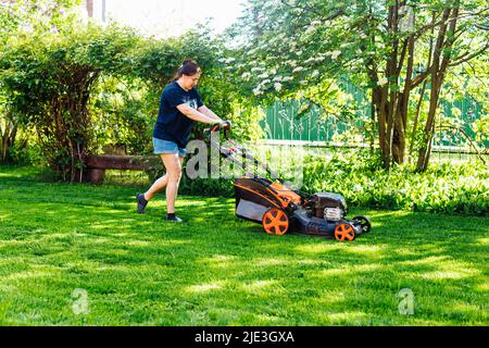 Donna dai capelli scuri in abiti casual con rasaerba a benzina o tagliaerba in giardino. Taglio di piante verdi fresche Foto Stock