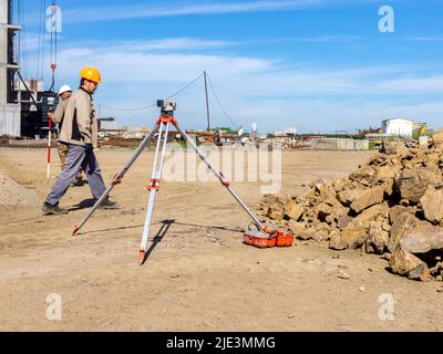 Kemerovo, Russia - Jule 02 2021. Un operatore di costruzione in un casco arancione cammina oltre un livello ottico in piedi su un cantiere vicino a un mucchio di Foto Stock