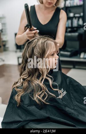 le mani di un parrucchiere con un ferro arricciante fanno un'acconciatura ad una ragazza curly in un salone di bellezza professionale Foto Stock