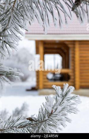 Primo piano di rami di abete ghiacciato ricoperti di neve in primo piano e veranda di casa di campagna in legno con tetto in tegole rosso sullo sfondo. Riposarsi Foto Stock