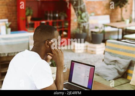 uomo d'affari multiculturale con laptop, telefono in caffetteria, tenere testa con mano, lavorare online come lavoratore autonomo Foto Stock