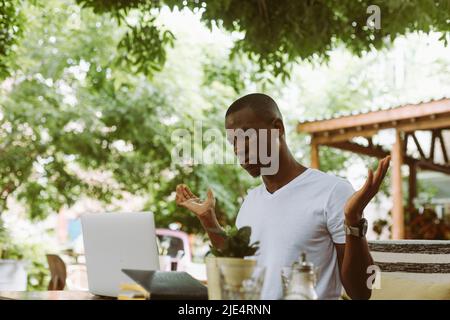 Sorpreso e confuso afroamericano uomo con computer portatile in caffè non sa cosa fare, alzando le mani in un caffè all'aperto Foto Stock