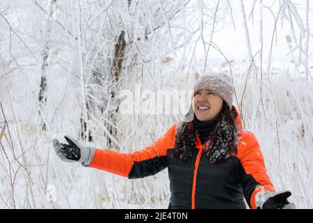 Donna felice di mezza età vestita calorosamente con la testa sorridente e le braccia distese a piedi in foresta nevosa con la neve caduta giù. Magico periodo invernale Foto Stock