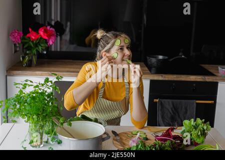 Donna bionda divertente e giocosa, casalinga con cetrioli a fette sulla pelle del viso come maschera naturale cuoco e mangiare in cucina Foto Stock