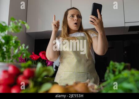 Grumo ispirato, più la donna bionda taglia in grembiule usando lo smartphone in cucina, indicizzandosi. Trova ricetta nell'app mobile Foto Stock