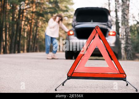 Primo piano sul triangolo cartello di avvertimento con la donna che chiede assistenza dopo aver scomposto con la sua auto sullo sfondo. Macchina rotta sulla strada Foto Stock
