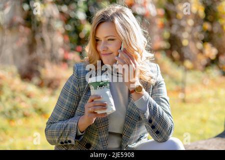 Giovane donna felice con lunghi capelli biondi in maglione e giacca sorride mentre parla al telefono, seduto nel parco della città sullo sfondo di alberi autunnali Foto Stock