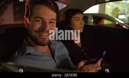 Uomo felice in auto dietro il sedile di notte dopo il lavoro ritratto sorridente tenendo il dispositivo smartphone. Due persone all'interno del taxi Foto Stock