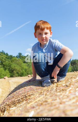 un ragazzo dai capelli rossi si siede sopra una pila di paglia dorata in un campo, un ragazzo su una pila di paglia di grano pungente, un ritratto di un ragazzo di sette anni Foto Stock