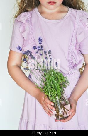 Ragazza giovane in un abito di lavanda pallido che tiene un vaso contenente fiori di lavanda appena raccolti su uno sfondo bianco Foto Stock
