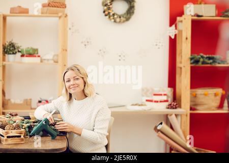 Ritratto di sorridente soddisfatto bella donna bionda in maglione bianco seduta a tavola in officina e di lavoro con materiali naturali per il Natale minaccia Foto Stock