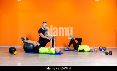 coppia senior che fa l'esercizio di sollevamento del peso che si posa in palestra isolato su sfondo arancione con personal trainer. persone mature fitness Foto Stock