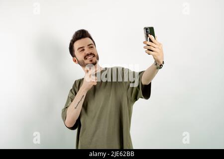 Allegro uomo caucasico indossare abiti casual posa isolato su sfondo bianco puntando dito indice sul telefono cellulare mentre prendere selfie con grande Foto Stock