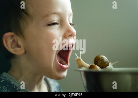 Il ragazzino aprì la bocca davanti alle lumache. Alimenti per bambini. Veganismo. Allevare i bambini e trattare gli animali Foto Stock