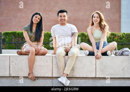 Amici multirazziali che guardano la macchina fotografica e sorridono mentre si siedono insieme all'aperto. Concetto di amicizia. Foto Stock