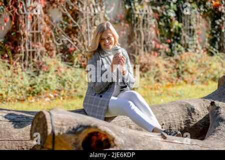 la bionda flirtatious è seduta sul tronco dell'albero nel parco della città, la donna è vestita in jeans bianchi e giacca a plaid, tenendo una tazza di cioccolata calda nelle mani Foto Stock