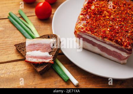 Pancetta piccante affumicata su un piatto. Fetta di carne di maiale affumicata su pane, pomodori ciliegini e cipolla verde su tavola di legno. Foto Stock