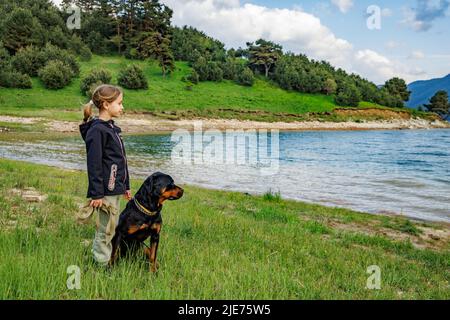 Bambina con guinzaglio in mani si alza vicino al suo grande cane della razza Rottweiler in verde prato erboso con vegetazione di montagna accanto a pulito piccolo Foto Stock