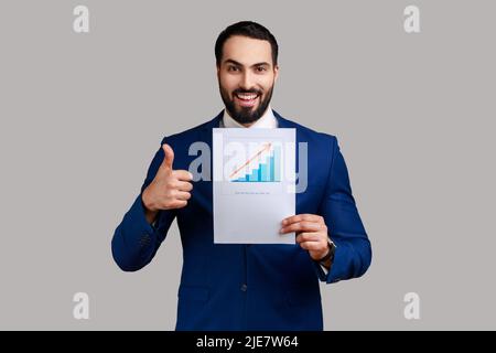 Bell'uomo d'affari barbuto in piedi mostrando il grafico della crescita aziendale, guardando la fotocamera, mostrando il pollice in su, indossando abito stile ufficiale. Studio interno girato isolato su sfondo grigio. Foto Stock