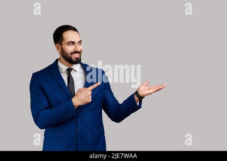 Uomo bearded che presenta lo spazio della copia sul suo palmo e che osserva sorridente, mostrando il posto vuoto per il testo promozionale, indossando il vestito di stile ufficiale. Studio interno girato isolato su sfondo grigio. Foto Stock