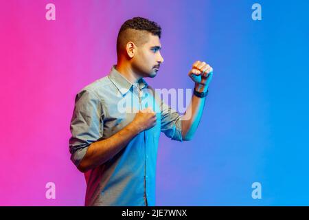 Ritratto laterale di giovane adulto arrabbiato aggressivo uomo in camicia punzonatura, pugilato con pugni clenched, essendo pronto a combattere. Studio interno girato isolato su colorato sfondo di luce al neon. Foto Stock