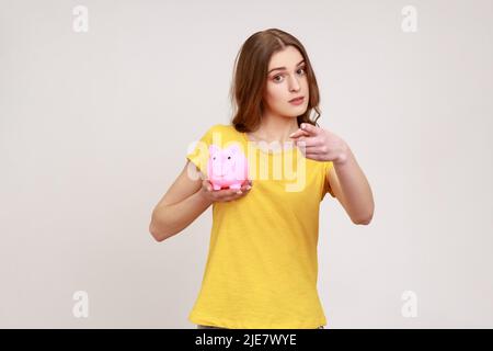 Bella donna di giovane età in giallo T-shirt investire in bitcoins, tenendo la banca piggy e puntando su di voi con il dito, guardando la macchina fotografica con il sorriso. Studio interno girato isolato su sfondo grigio. Foto Stock