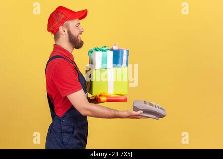 Vista laterale del corriere sorridente in tute e T-shirt rossa che tiene le scatole avvolte, caming al cliente con ordine, dando pos contactless terminale di pagamento. Studio interno girato isolato su sfondo giallo. Foto Stock