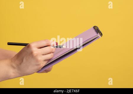 Profilo Vista laterale primo piano della mano donna scrittura in blocco note con penna o matita, facendo a di list. Studio interno girato isolato su sfondo giallo. Foto Stock