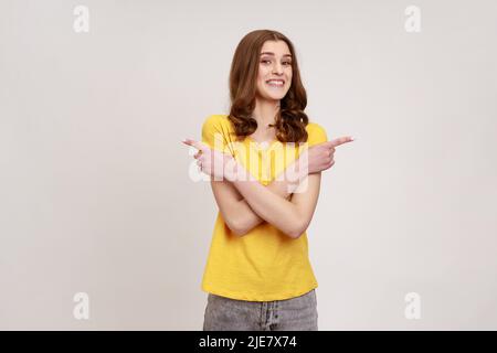 Ritratto di positivo estremamente felice donna di giovane età in giallo T-shirt in piedi con le mani incrociate e puntando allo spazio di copia su entrambi i lati. Studio interno girato isolato su sfondo grigio. Foto Stock