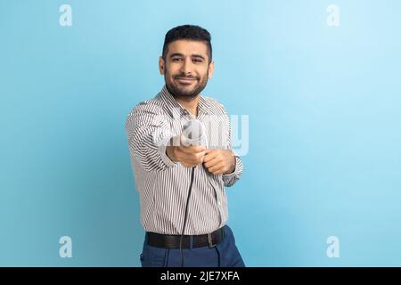 Ritratto di uomo d'affari sorridente e attraente con barba in piedi che offre microfono, giornalista che prende l'intervista, indossando una camicia a righe. Studio interno girato isolato su sfondo blu. Foto Stock
