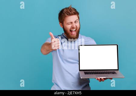 Ritratto di uomo con bearded positivo che tiene il laptop con schermo vuoto, winking, soddisfatto con l'applicazione del calcolatore, guardando la fotocamera, mostrando il pollice in su. Studio interno girato isolato su sfondo blu. Foto Stock