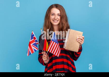 Studentessa positiva indossando un pullover casual a righe, con bandiere di libri di tedesco, USA e Regno Unito, imparare le lingue, educare all'estero. Studio interno girato isolato su sfondo blu. Foto Stock