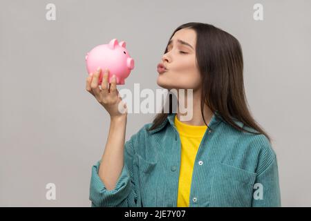 Ritratto di felice donna soddisfatto tenendo Piggy banca in mani con pout labbra, baciare, risparmio di denaro, indossare casual stile giacca. Studio interno girato isolato su sfondo grigio. Foto Stock