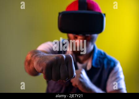 Hipster uomo in occhiali di realtà virtuale sulla testa giocando il gioco di combattimento, tenendo i pugni clenched fino alla macchina fotografica, pronto a boxe. Studio interno girato isolato su colorato sfondo di luce al neon. Foto Stock