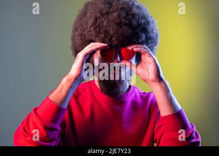 Ritratto dell'uomo hipster con acconciatura afro che copre gli occhi con piccoli cuori rossi, esprimendo sentimenti romantici, indossando una felpa rossa. Studio interno girato isolato su colorato sfondo di luce al neon. Foto Stock