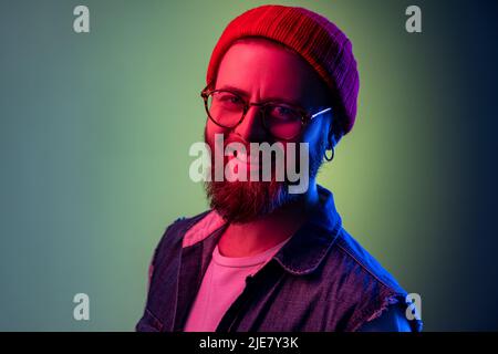 Estremamente felice hipster uomo guardante la macchina fotografica con il sorriso toothy, ottimismo, successo, indossando berretto rosso di beanie e gilet di denim. Studio interno girato isolato su colorato sfondo di luce al neon. Foto Stock