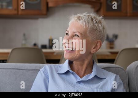 Donna anziana felice che staring da parte seduto sul divano a casa Foto Stock