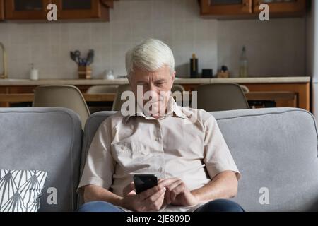 Uomo singolo dai capelli grigi seduto sul divano utilizzando lo smartphone Foto Stock