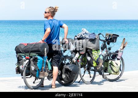 Un uomo si riposa e si alza reggendo due biciclette mentre guarda verso il mare. Le biciclette sono molto cariche di equipaggiamento per una vacanza o un tour in bikepacking Foto Stock