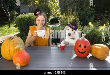 giovane donna in abito arancione con cane in cappelli strega sono seduti a tavola in giardino, bere caffè, riposare. tavolo è decorato con pumpki arancione Foto Stock