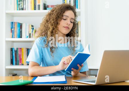 Graziosa studentessa caucasica che legge un libro di riferimento al banco dell'università Foto Stock