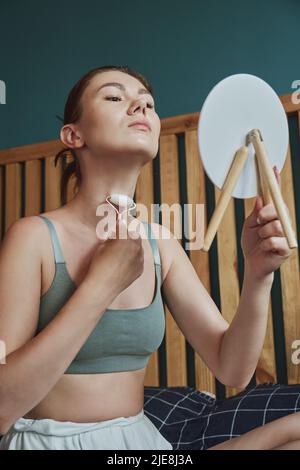 Giovane donna che usa il rullo facciale della giada per il massaggio del viso seduto sul letto in camera da letto guardando nello specchio Foto Stock