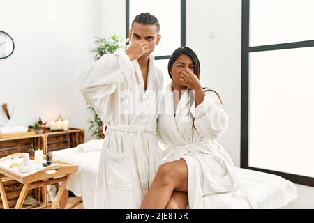 Giovane coppia latina che indossa un asciugamano in piedi al centro di bellezza che odora qualcosa di stinky e disgustoso, odore intollerabile, tenendo il respiro con le dita sopra Foto Stock