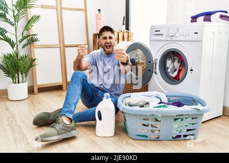 Giovane uomo ispanico che mette la lavanderia sporca in lavatrice arrabbiato e pazzo che alza i pugni frustrati e furiosi mentre gridano con rabbia. Rabbia e un Foto Stock