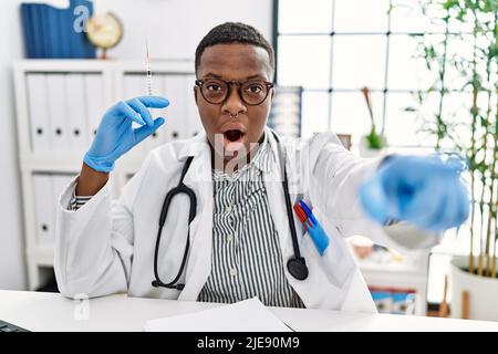 Giovane medico africano che tiene la siringa all'ospedale puntando con il dito sorpreso davanti, bocca aperta stupito espressione, qualcosa sul fronte Foto Stock