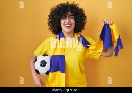 Giovane donna brunetta con capelli ricci football hooligan tenere palla sorridendo e ridendo forte fuori perché divertente scherzo pazzo. Foto Stock