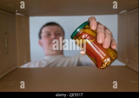 Un uomo toglie il cibo da una scatola di cartone. Verdure in scatola. Vasetto di vetro con peperoncino estratto dalla confezione della donazione. Vista dal basso. Vista interna. Primo piano Foto Stock