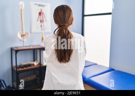 Giovane donna bruna che lavora alla clinica di recupero del dolore in piedi indietro guardando via con le braccia incrociate Foto Stock
