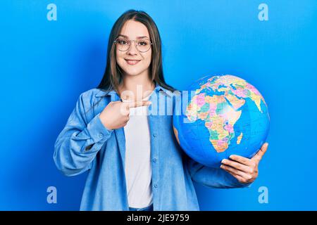 Giovane ragazza ispanica che tiene la sfera del mondo sorridendo felice indicando con mano e dito Foto Stock